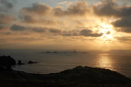 This landscape photograph captures the coast at sunset, focusing on the Longships lighthouse in the waters off Sennen Cove near Land's End. The image was taken in the evening during the autumn season, as indicated by the soft, warm hues of the sinking sun and the partially clouded sky. Sunlight reflects off the sea, casting a golden glow across the coastline and surrounding headland. The distinctive Longships lighthouse stands among rocky outcrops in the distance, silhouetted against the bright horizon and illuminated by the last light of day. The rugged terrain of the Cornish coast and the historic shipping landmark together create an atmospheric representation of autumn evenings at Land's End.